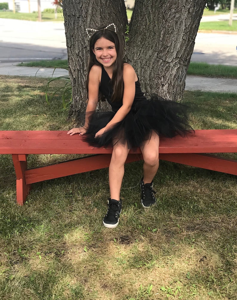 May include: A young girl wearing a black tulle tutu skirt and a black tank top with a cat ear headband. She is sitting on a red wooden bench with her legs crossed and smiling at the camera.