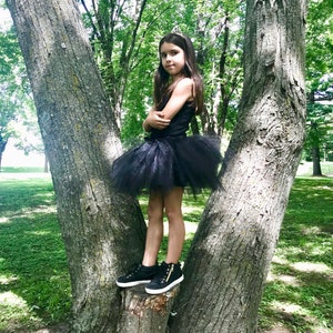 May include: A young girl wearing a black tulle skirt and black sneakers stands on a tree trunk in a forest setting.