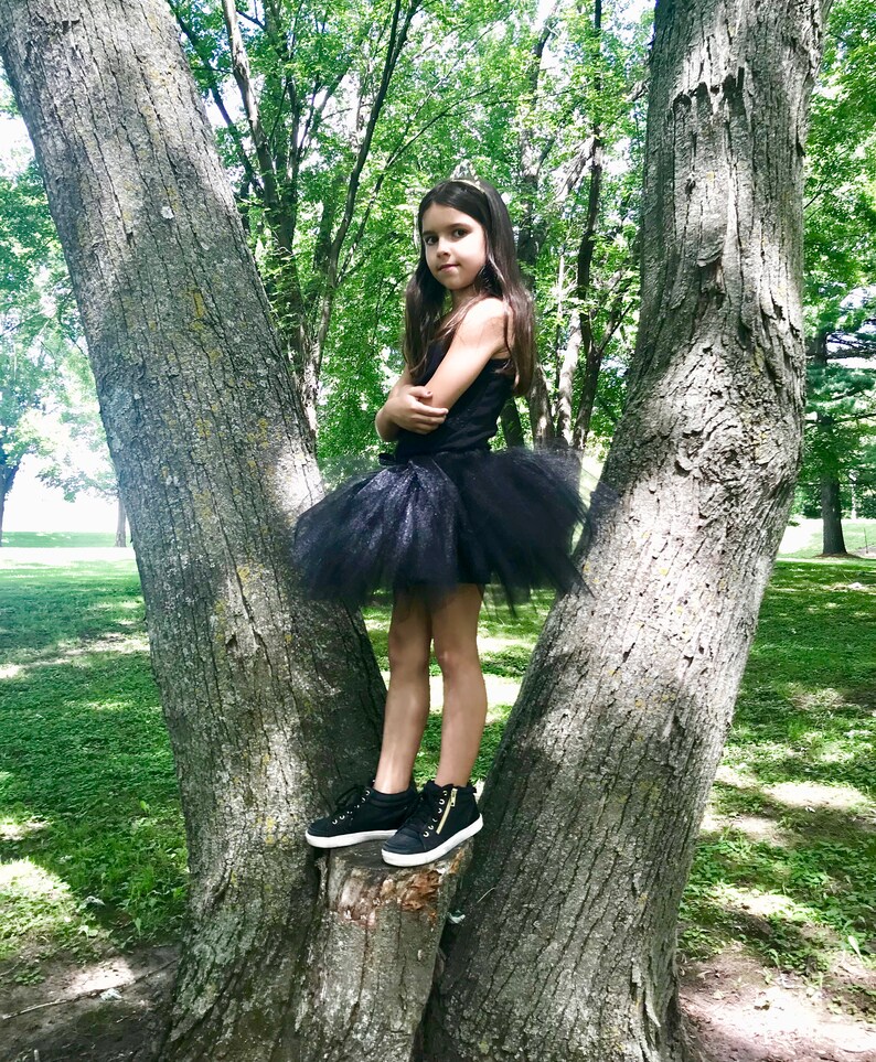 May include: A young girl wearing a black tulle skirt and black sneakers stands on a tree trunk in a forest setting.
