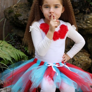 May include: A young girl wearing a white long-sleeved shirt with a red flower embellishment, a red and blue tulle tutu skirt, and red leg warmers. She is holding her finger to her lips, as if to say 'shhh'.