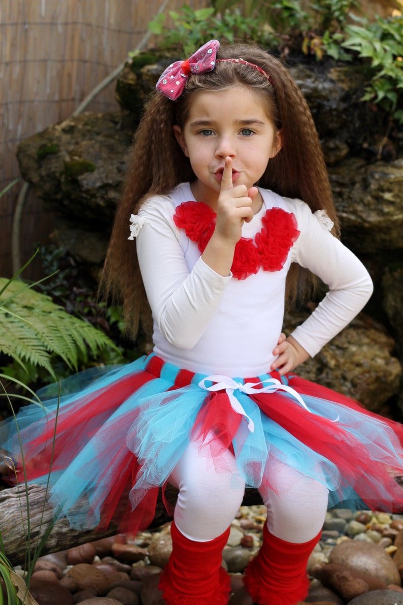 May include: A young girl wearing a white long-sleeved shirt with a red flower embellishment, a red and blue tulle tutu skirt, and red leg warmers. She is holding her finger to her lips, as if to say 'shhh'.