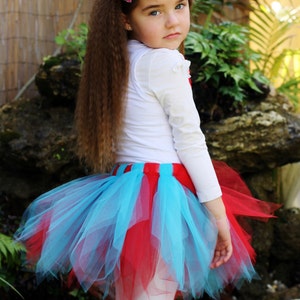 May include: A young girl wearing a white long-sleeved shirt, a red and blue tulle skirt, and a pink polka dot bow in her hair. She is standing in front of a rock and a wooden fence.