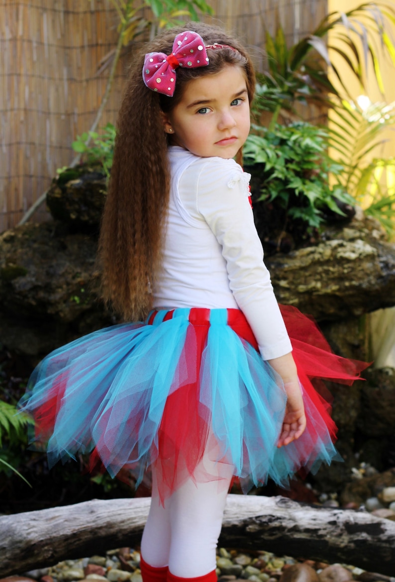 May include: A young girl wearing a white long-sleeved shirt, a red and blue tulle skirt, and a pink polka dot bow in her hair. She is standing in front of a rock and a wooden fence.
