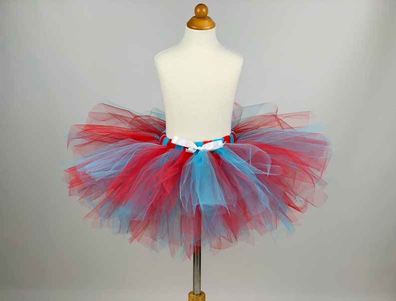 May include: A red and blue tulle tutu with a white satin ribbon. The tutu is on a white mannequin.