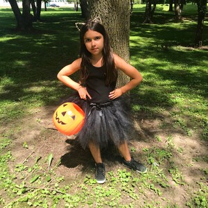 May include: A young girl dressed as a black cat for Halloween, wearing a black tulle skirt, a black tank top, and a black headband with cat ears. She is holding an orange plastic pumpkin bucket and standing in a grassy area with trees in the background.