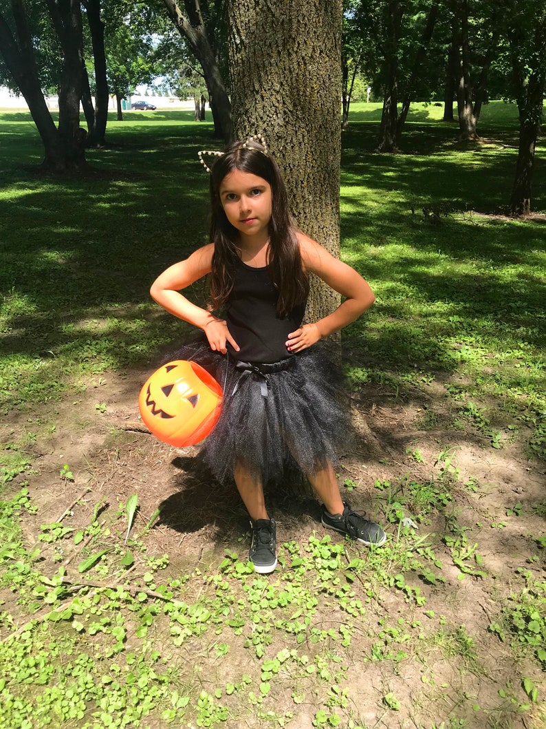 May include: A young girl dressed as a black cat for Halloween, wearing a black tulle skirt, a black tank top, and a black headband with cat ears. She is holding an orange plastic pumpkin bucket and standing in a grassy area with trees in the background.