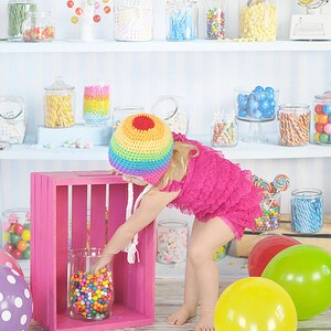 May include: A young girl wearing a rainbow crochet hat and a pink dress reaches into a glass jar filled with colorful candy. The jar is sitting on a pink wooden crate. There are balloons in the background.