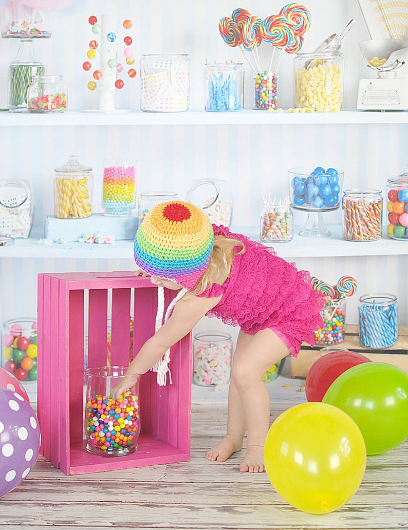 May include: A young girl wearing a rainbow crochet hat and a pink dress reaches into a glass jar filled with colorful candy. The jar is sitting on a pink wooden crate. There are balloons in the background.