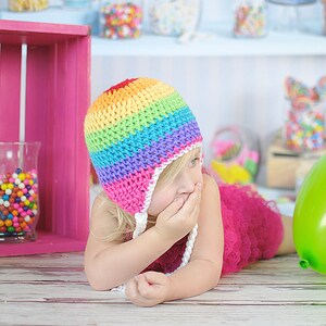 May include: A young girl wearing a rainbow crocheted hat with a white brim and a pink tulle dress. She is covering her mouth with her hand and looking to the side. A green balloon is on the floor next to her.