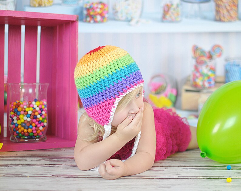May include: A young girl wearing a rainbow crocheted hat with a white brim and a pink tulle dress. She is covering her mouth with her hand and looking to the side. A green balloon is on the floor next to her.