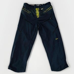 May include: Dark navy blue Oakley pants with yellow accents and multiple zippered pockets. The pants have a relaxed fit and a small Oakley logo on the lower leg. The pants are made of a durable, water-resistant material.
