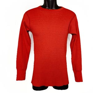 Könnte beinhalten: Ein langärmeliges, rotes Thermoshirt mit Waffelstrickstruktur. Das Shirt hat einen Rundhalsausschnitt und gerippte Bündchen. Das Kleidungsstück wird auf einer schwarzen Schaufensterpuppe vor weißem Hintergrund präsentiert. Das Shirt ist einfarbig.