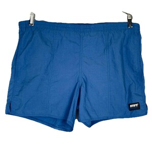 May include: Bright blue athletic shorts featuring an elastic waistband and vertical seam detailing. A small black rectangular patch is located on the lower right leg, displaying the white Nike logo and text. The shorts have a smooth, lightweight finish.