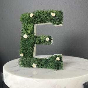 以下が含まれることがあります： 緑色の苔と小さな白い花で覆われた装飾的な文字「E」。文字は白い素材でできており、白い大理石の表面に置かれています。背景は濃い灰色です。