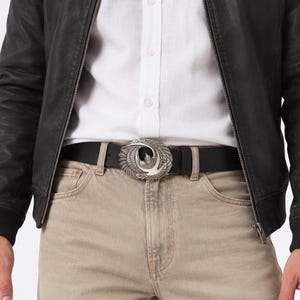 May include: A black leather belt featuring a silver-toned oval buckle with an embossed eagle design. The belt is worn with a white button-down shirt, beige trousers, and a black leather jacket, showcasing the detailed metalwork of the bird motif.