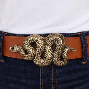 May include: A brown leather belt with a brass snake buckle. The snake buckle is intricately detailed with a textured pattern. The belt is worn with blue denim jeans and a white shirt.
