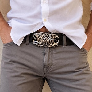 May include: A black leather belt featuring a metallic silver-toned buckle shaped like two mirrored lion heads facing outward. The buckle has intricate detailing on the manes and facial features, secured onto a smooth black strap with a standard prong.
