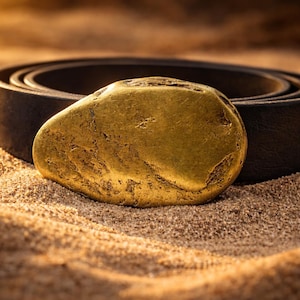 May include: A golden, irregularly shaped object rests on a bed of sand, with a dark brown leather belt in the background. The object has a textured surface and appears to be made of metal. The lighting suggests a warm, sunny environment.