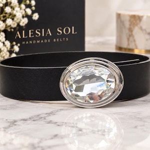 May include: A black belt featuring a large, oval-shaped, faceted clear crystal buckle set in a silver-toned metal frame. The belt strap is smooth and solid black. The text ALESIA SOL HANDMADE BELTS is visible on a black box in the background.