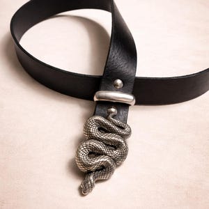May include: Black leather belt featuring a silver-coloured snake buckle. The belt has a classic design with a single loop and a metal buckle. The snake buckle is intricately detailed, adding a unique touch to the accessory.