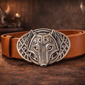 May include: A tan leather belt featuring a large, oval-shaped silver-toned metal buckle. The buckle displays a detailed, embossed wolf head design with intricate geometric and knotwork patterns surrounding the animal's face in a metallic finish.