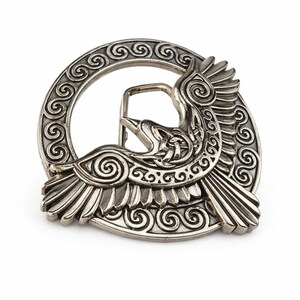 May include: A silver-colored belt buckle featuring a detailed design of a bird in flight. The bird is surrounded by a circular frame with intricate spiral patterns. The buckle has a classic pin and loop closure.