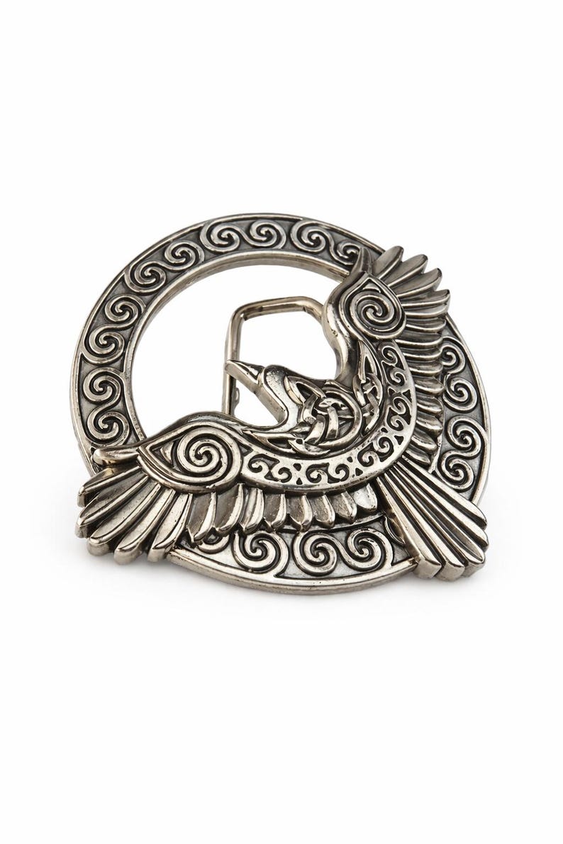 May include: A silver-colored belt buckle featuring a detailed design of a bird in flight. The bird is surrounded by a circular frame with intricate spiral patterns. The buckle has a classic pin and loop closure.
