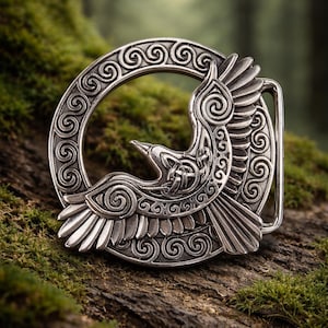 May include: A silver-colored belt buckle featuring a detailed design of a bird in flight, set against a circular frame with intricate scrollwork. The buckle is displayed on a mossy, textured surface, suggesting a nature-inspired theme.