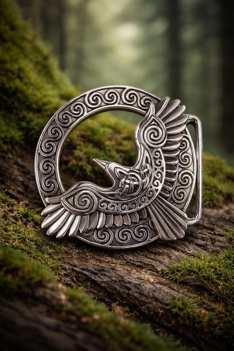 May include: A silver-colored belt buckle featuring a detailed design of a bird in flight, set against a circular frame with intricate scrollwork. The buckle is displayed on a mossy, textured surface, suggesting a nature-inspired theme.