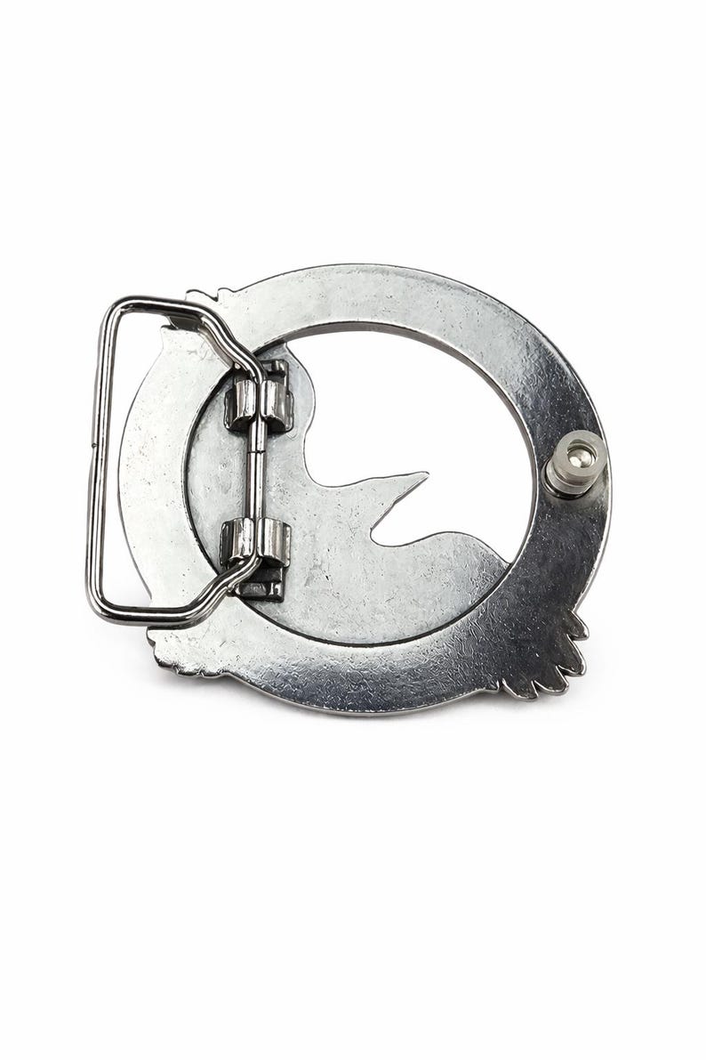 May include: A silver-colored metal belt buckle with a bird design. The buckle has a circular frame with a bird silhouette in the center. The buckle has a rectangular bar and a pin for securing the belt.