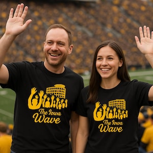 May include: A black short-sleeved t-shirt featuring a yellow graphic print of a stadium and a crowd with raised hands. The text "The Iowa Wave" is printed in yellow below the graphic. The design is centered on the chest of the garment.
