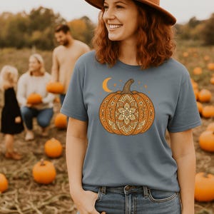 May include: A woman wearing a blue t-shirt with a detailed orange pumpkin graphic, a brown hat, and blue jeans. The pumpkin design includes a floral mandala pattern, a crescent moon, and stars. The background shows a pumpkin patch.