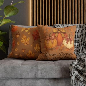 Può includere: Due cuscini decorativi con disegni a tema autunnale. Un cuscino presenta un motivo di foglie e ghiande nei toni dell'arancione e del giallo su sfondo marrone. L'altro mostra zucche con fiocchi e il testo "Bows, Leaves, & Pumpkins."