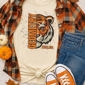 May include: Cream t-shirt featuring a tiger graphic and the word "GEOMETRY" in orange. The outfit includes a plaid shirt, blue jeans, and orange trainers. Autumnal decorations include pumpkins and leaves.