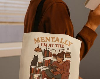 May include: Cream-colored tote bag with the text "Mentally I'm at the Bookstore." The graphic depicts a person reading in a chair, a black cat, and bookshelves. The bag has a black strap and is being held by a person.