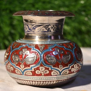 May include: A metallic vase featuring a wide, flared rim and a bulbous body. The surface is decorated with intricate engraved patterns filled with vibrant red and light blue enamel, showcasing floral and leaf motifs across the exterior.
