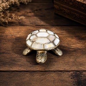 Peut inclure: Une figurine de tortue décorative dotée d'un corps en métal doré et d'une carapace incrustée de tuiles hexagonales en nacre blanche irisée. La tortue présente une finition polie et lisse sur la tête et les pattes, avec un motif de carapace détaillé.
