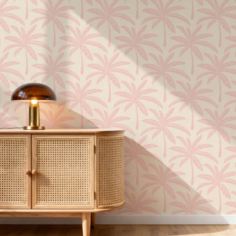 Puede incluir: Un mueble de madera clara con dos puertas de textura de rejilla tejida y pomos redondos de madera. Una l&aacute;mpara de mesa con pantalla de c&uacute;pula de cristal oscuro y base dorada se sit&uacute;a encima. El fondo es papel pintado con un patr&oacute;n de hojas de palma rosas.