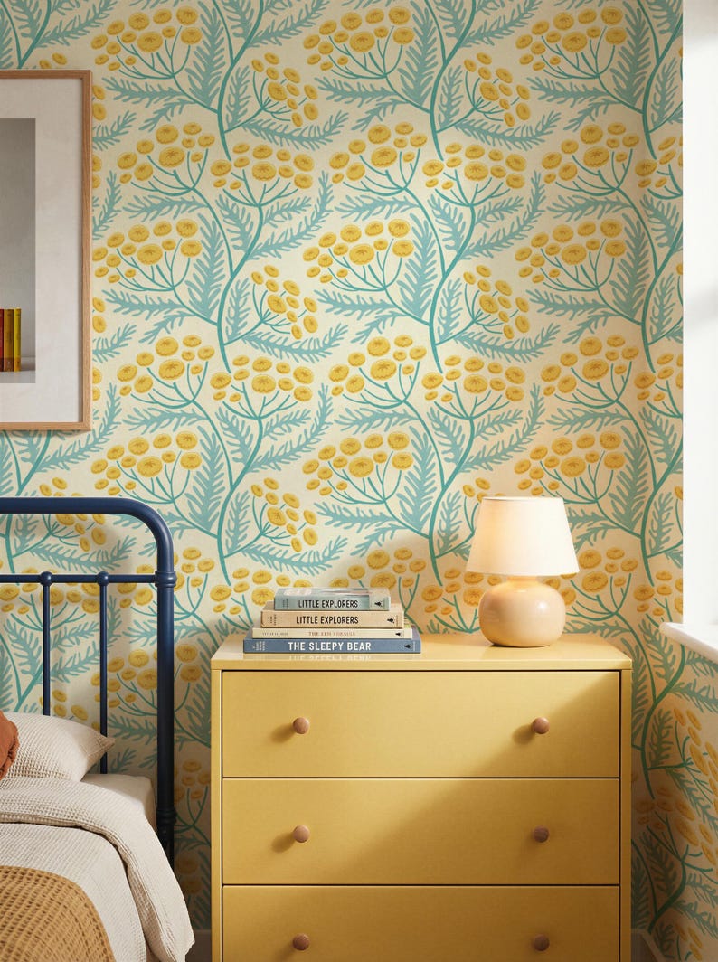 Blue and yellow cottagecore wildflower wallpaper. The flowers are hand drawn in a folk art botanical style on a cream background. Styled in a bedroom with a yellow dresser, a blue metal headboard, and a framed picture on the wall.