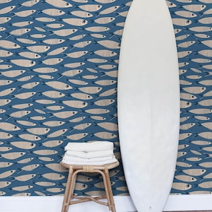 Puede incluir: Una tabla de surf blanca se apoya verticalmente junto a un pequeño taburete de madera con toallas blancas dobladas encima. El fondo es un papel pintado con un patrón repetitivo de peces beige con contornos oscuros sobre un fondo azul.