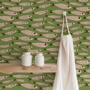 May include: A green wallpaper featuring a repeating pattern of beige fish with dark brown outlines and eyes. A wooden shelf holds two white ceramic jars, and a white apron hangs from a hook on the right side of the wall surface.
