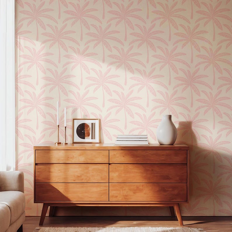 Puede incluir: Papel pintado con un patr&oacute;n repetitivo de siluetas de palmeras de color rosa claro sobre un fondo crema. El dise&ntilde;o cubre la pared detr&aacute;s de una c&oacute;moda de madera, dos candelabros altos y delgados, un cuadro enmarcado y un jarr&oacute;n blanco.