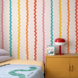 Rainbow wiggle stripe wallpaper featuring red orange yellow blue and pink wavy Ric Rac lines on a warm white background.  Styled in a modern trendy kids bedroom with yellow metal locker, red lamp and child's bed.