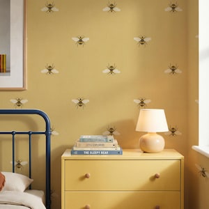 Op de afbeelding: Mosterdgeel behang met een herhalend patroon van kleine, donkerbruine en witte bijen. De muur is versierd met een ingelijste afbeelding, een gele ladekast met drie lades, een kleine lamp en een stapel boeken genaamd Little Explorers en The Sleepy Bear.