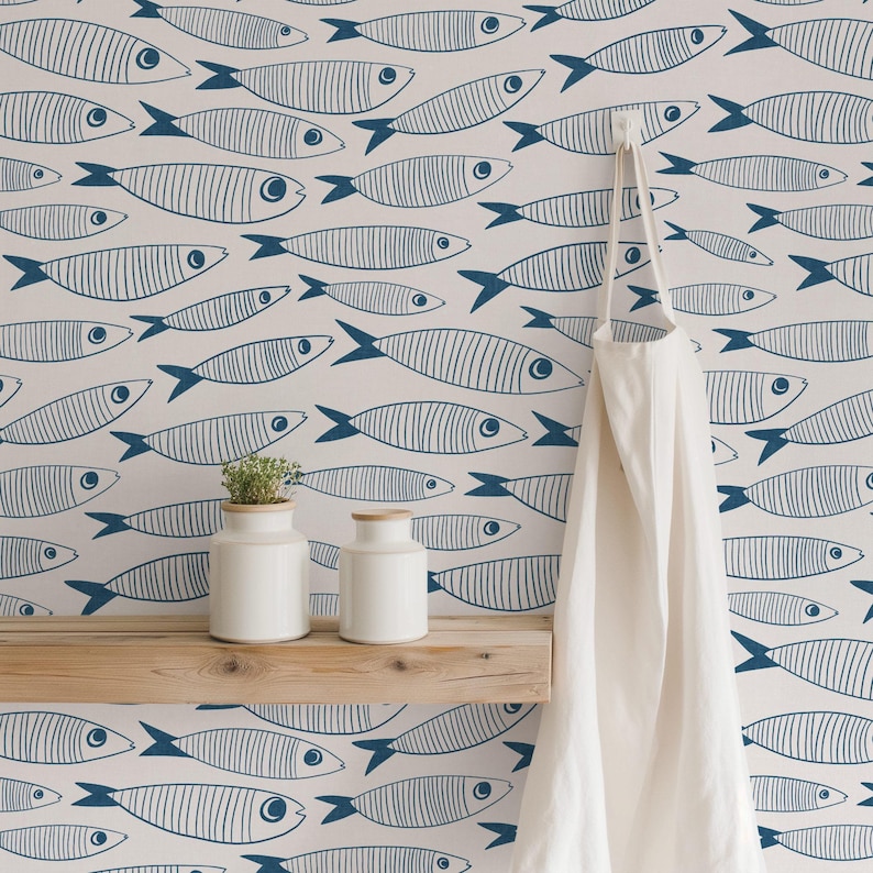 Blue and white sardine fish print wallpaper in a playful coastal style, shown in a kitchen nook with a wooden shelf, ceramic jars, and apron hanging for scale.