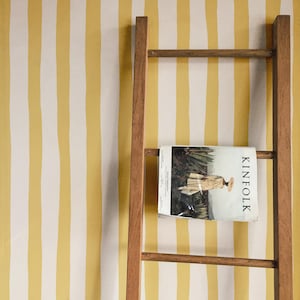 Pode incluir: Uma escada de madeira encostada a uma parede com papel de parede de riscas verticais amarelas e brancas. Um exemplar da revista Kinfolk está pousado no degrau central da escada, exibindo uma capa com uma pessoa de chapéu num deserto.