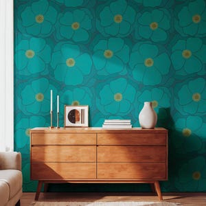 May include: A wooden chest of drawers with six compartments stands against a wall covered in teal wallpaper featuring a repeating pattern of large, dark-outlined flowers with yellow centres. On the unit sit two tall candlesticks, a framed print, and a white vase.