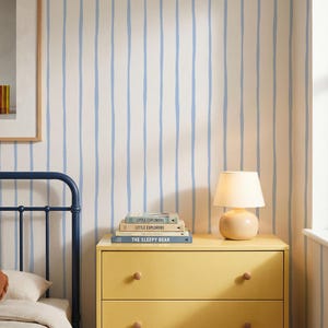 Può includere: Carta da parati con strisce verticali color azzurro disegnate a mano su sfondo crema. Il motivo si ripete sulla parete, creando un design lineare coerente che copre la superficie dietro una cassettiera gialla e una struttura letto blu scuro.