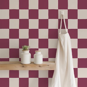 Op de afbeelding: Een muur met een bordeauxrood en crèmekleurig dambordpatroon heeft een plank van licht hout met twee witte keramische potten, waarvan er één een kleine groene plant bevat. Een effen wit schort hangt aan een haak aan de muur.