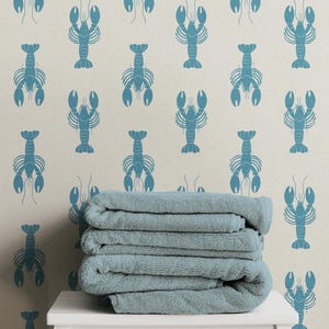 May include: A stack of three folded light blue towels resting on a white surface against a light beige wall featuring a repeating pattern of blue lobster illustrations arranged in vertical columns across the entire surface of the wall.